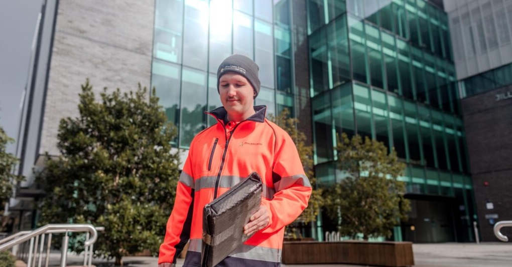 Male courier in orange high-visibility jacket delivering a package in front of modern building, Wollongong.